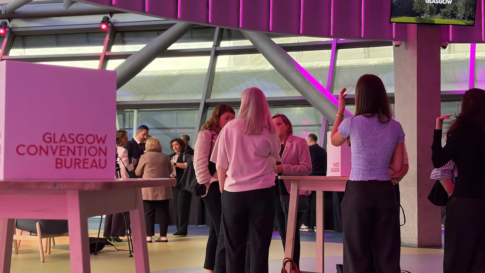 Several attendees gather around tables in a convention hall with pink signage for the Glasgow Convention Bureau.