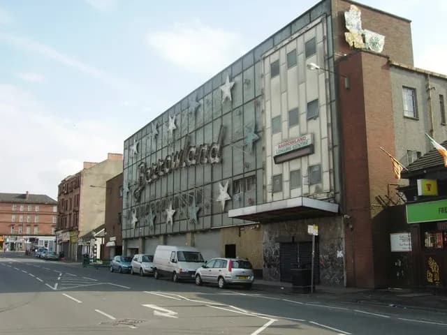 The external facade of the Barrowland Ballroom in Glasgow, a large building adorned with stars and its name in prominent lettering, facing a street with parked cars and other buildings in the distance under a blue sky.