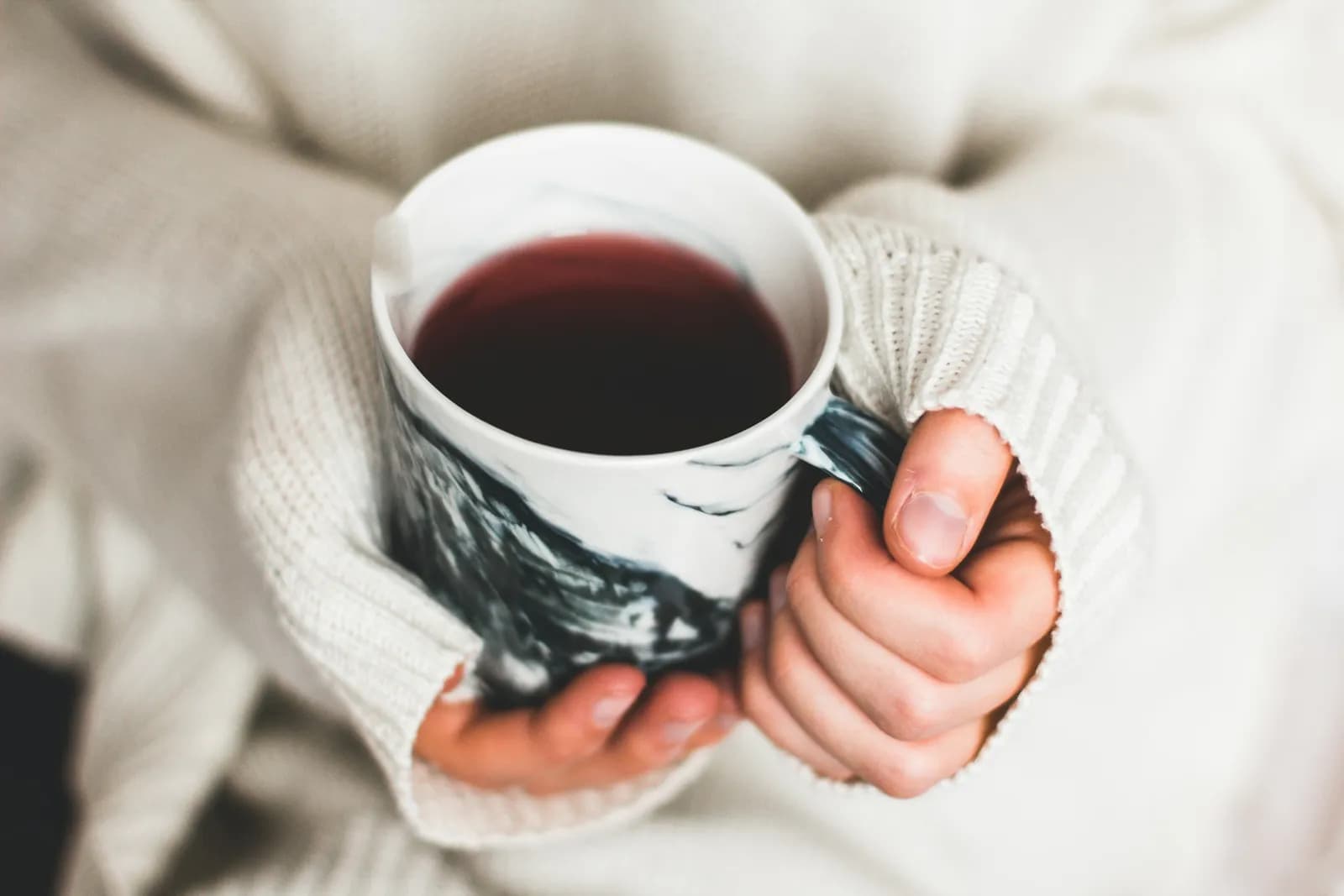 A person wearing a white knit sweater holds a white marble-patterned mug filled with a dark red beverage.